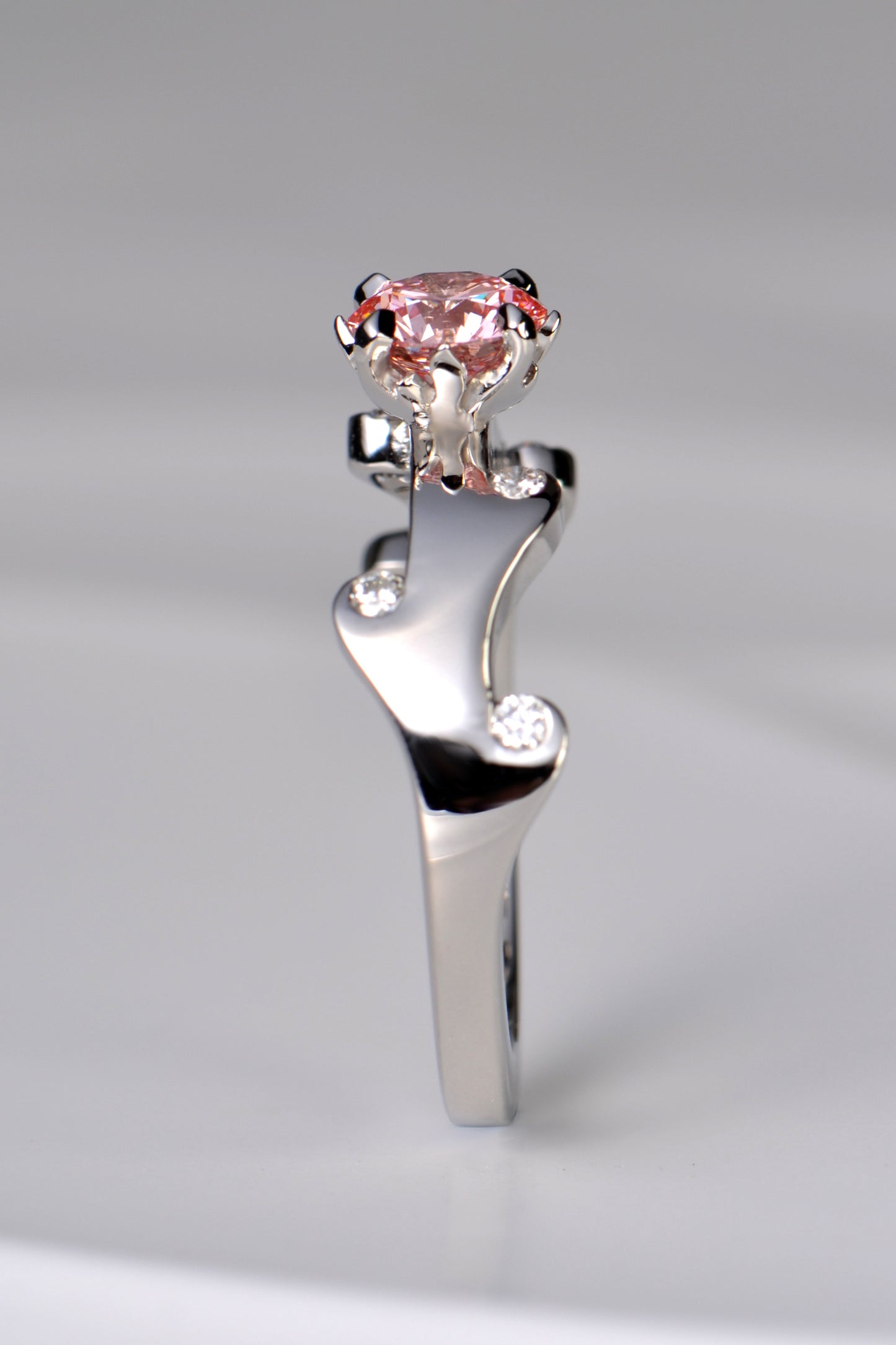 A stunning platinum engagement ring that is a unique design inspired by fire. The band waves in curving flames that hold small white lab diamonds and the main central round pink lab diamond is held by claws that are shaped into tiny flames.