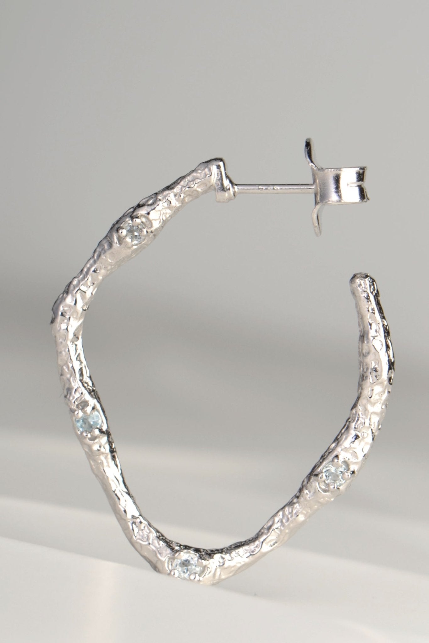 large silver hoop earrings with molton lava texture set with four very small pale blue topaz gemstones on one side by jewellery designer Jorge Revilla