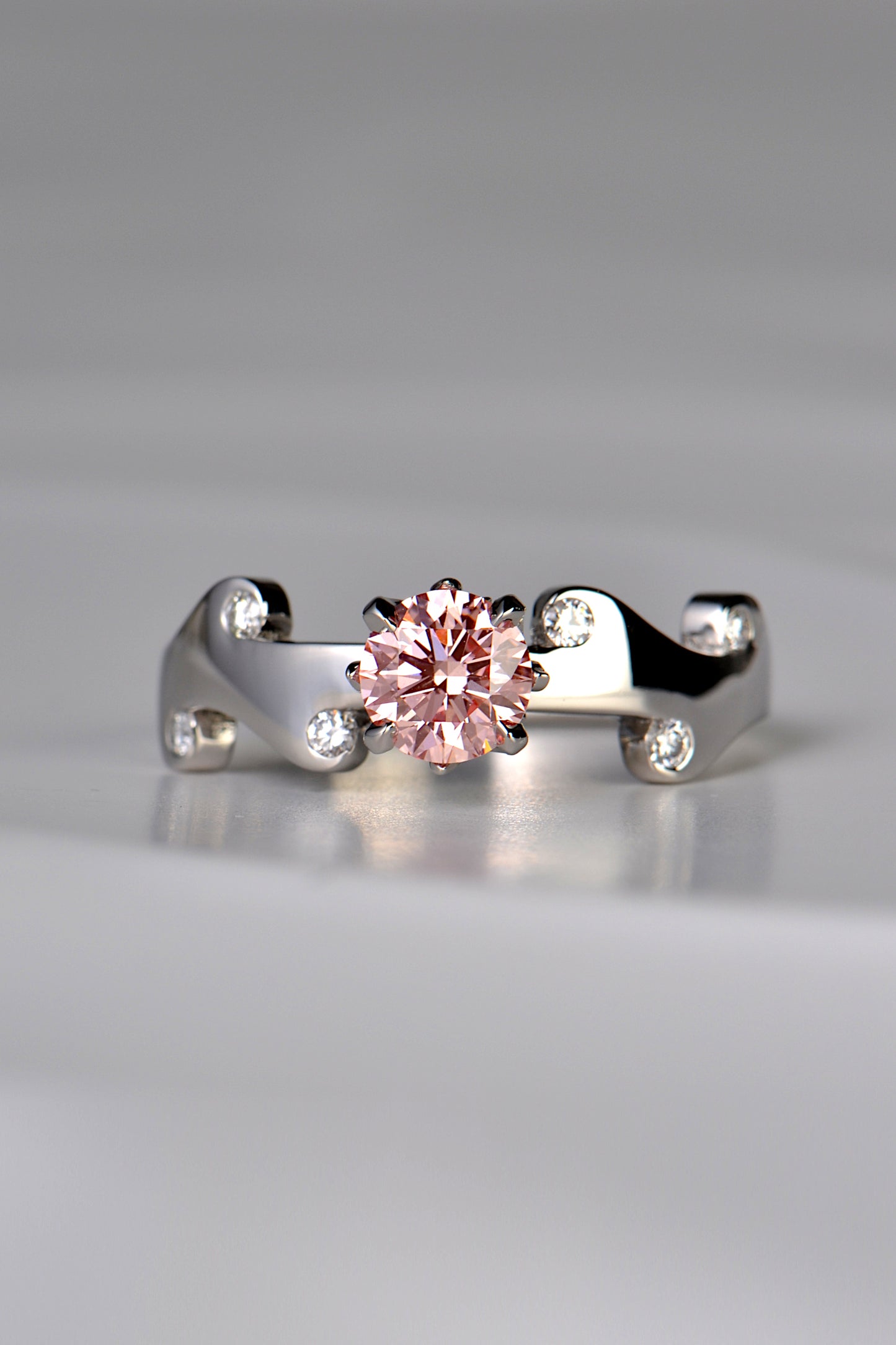 Large round pink lab diamond set in the middle of a platinum wavy ring with 6 small round white diamonds scattered across the front half of the band. This unique design by Christine Sadler is inspired by fire and the pink lab diamond is held by claws that are shaped into tiny flames.