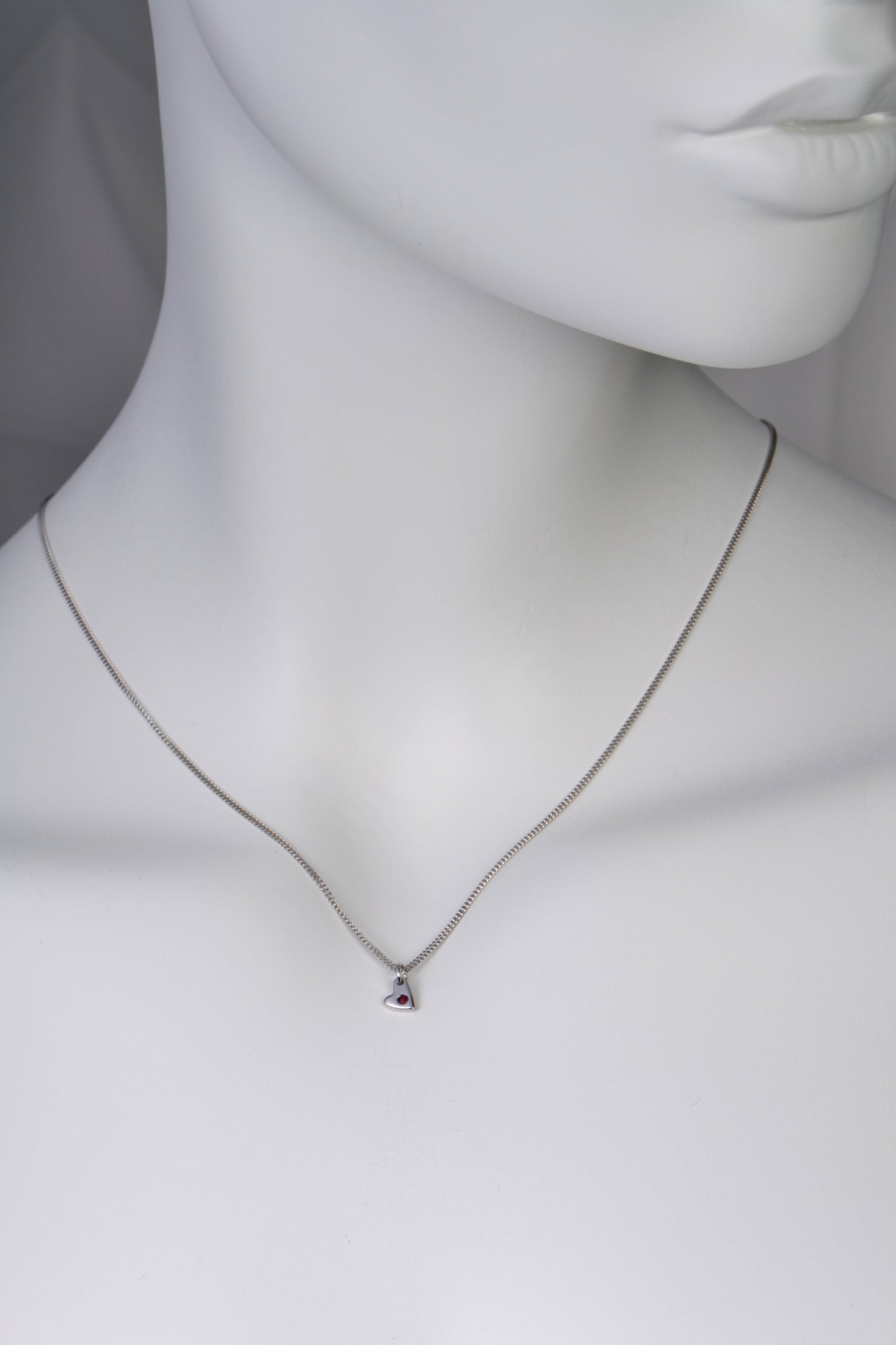 Small polished white gold heart necklace with a ruby displayed on white mannequin to show size and scale on a 40cm long chain.