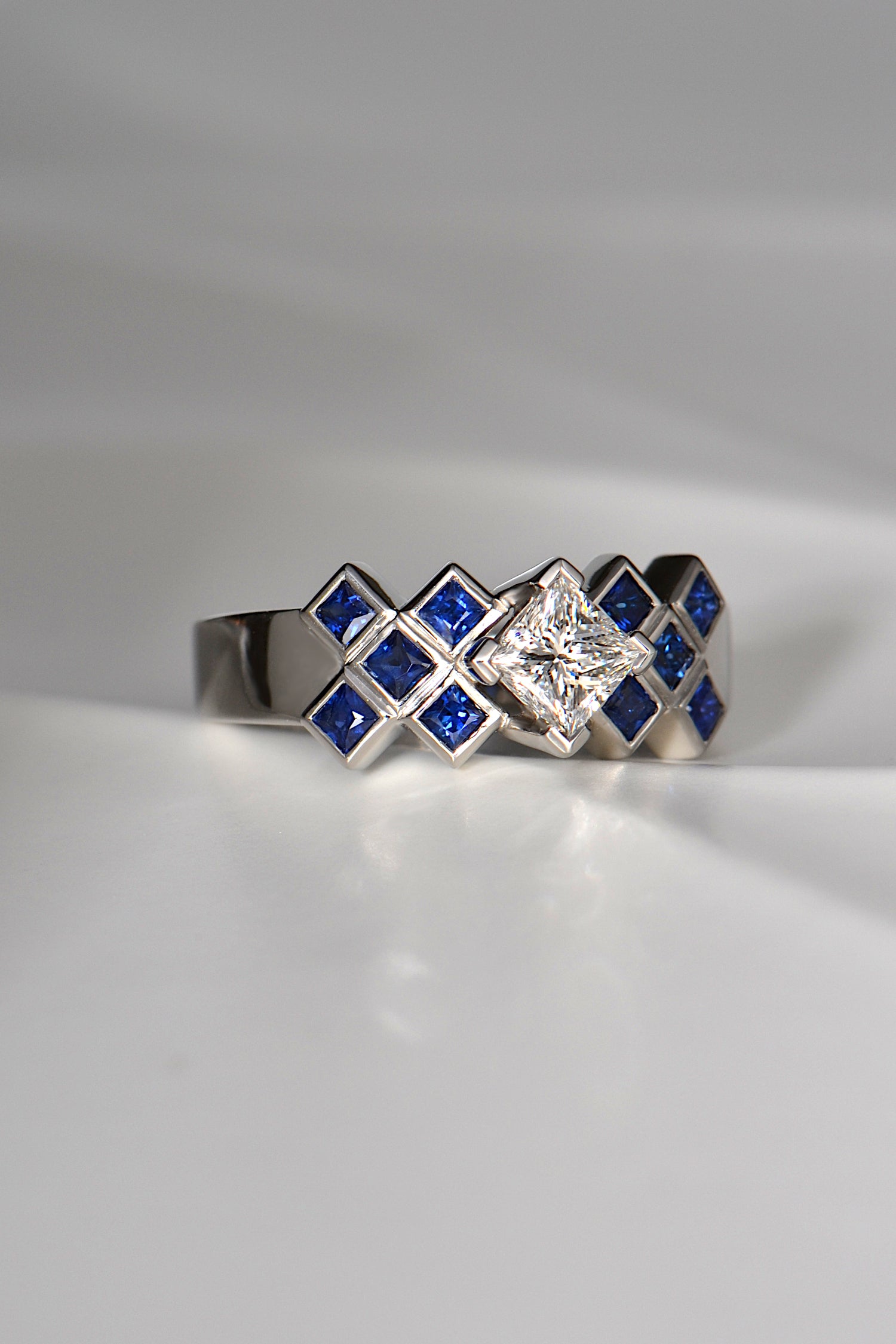 A large centre princess cut white diamond is set above a sea of square cut blue sapphires in a platinum engagement ring. There are ten sapphires in total, five on either side of the princess cut central diamond.
