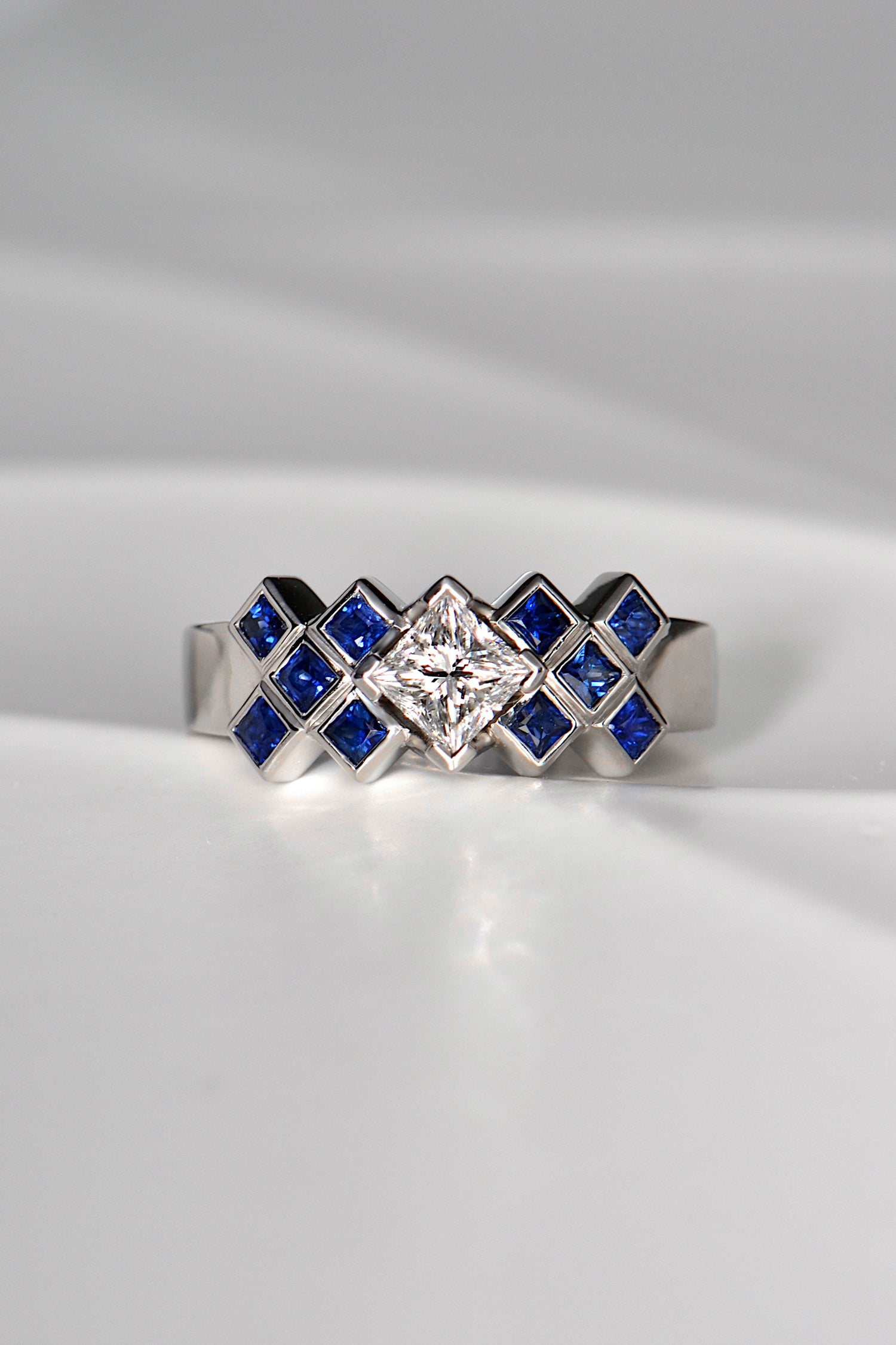 Princess cut diamond ring set with square blue sapphires in a tartan pattern. The ring is made of platinum and is a unique copyrighted design. 