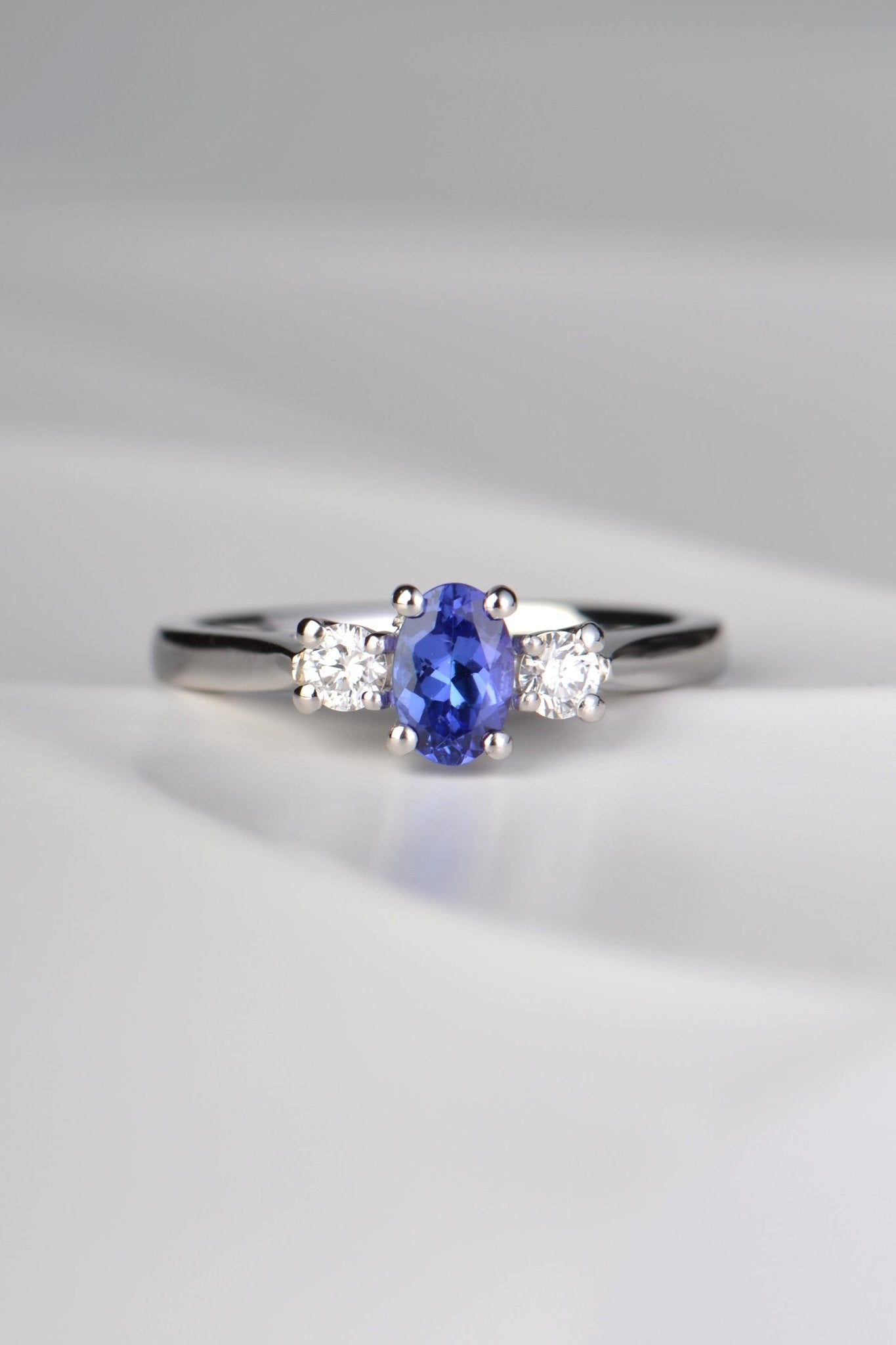 stunning oval cut intense blue tanzanite engagement ring set with two round diamonds, one on either side of the tanzanite. Each stone is held by four claws and it is made in platinum.
