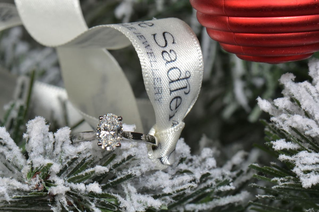 an oval lab diamond engagement ring on a Christmas tree branch with a red bauble decoration