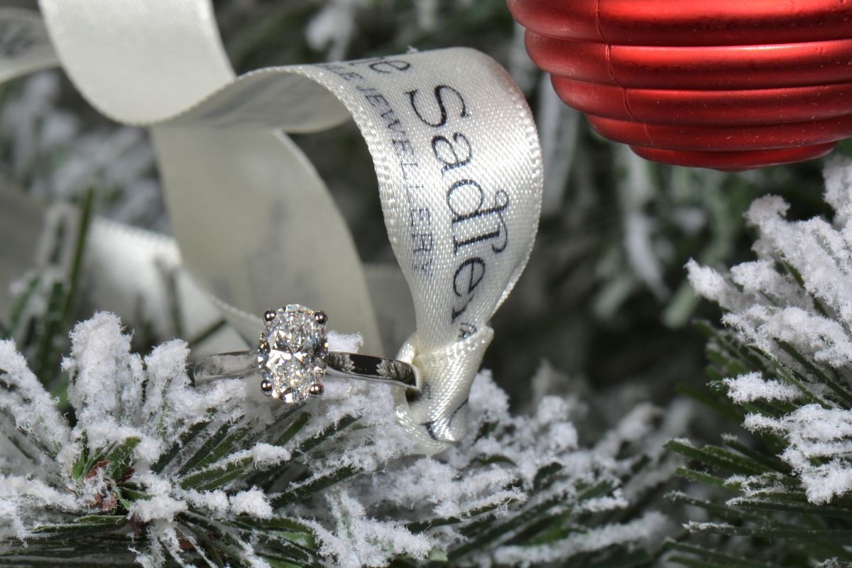 an oval lab diamond engagement ring on a Christmas tree branch with a red bauble decoration