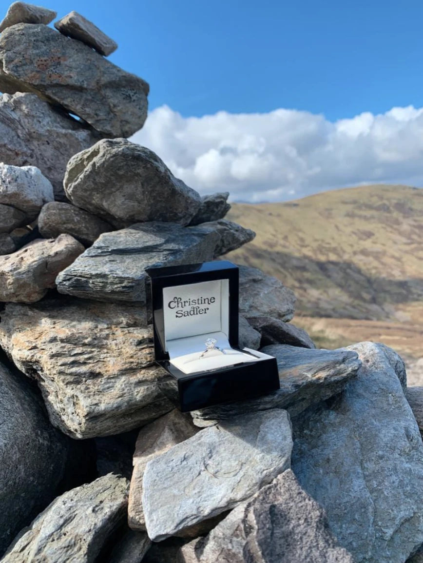 Open jewellery box with an engagement ring from Christine Sadler on a stack of rocks against a scenic Scottish background