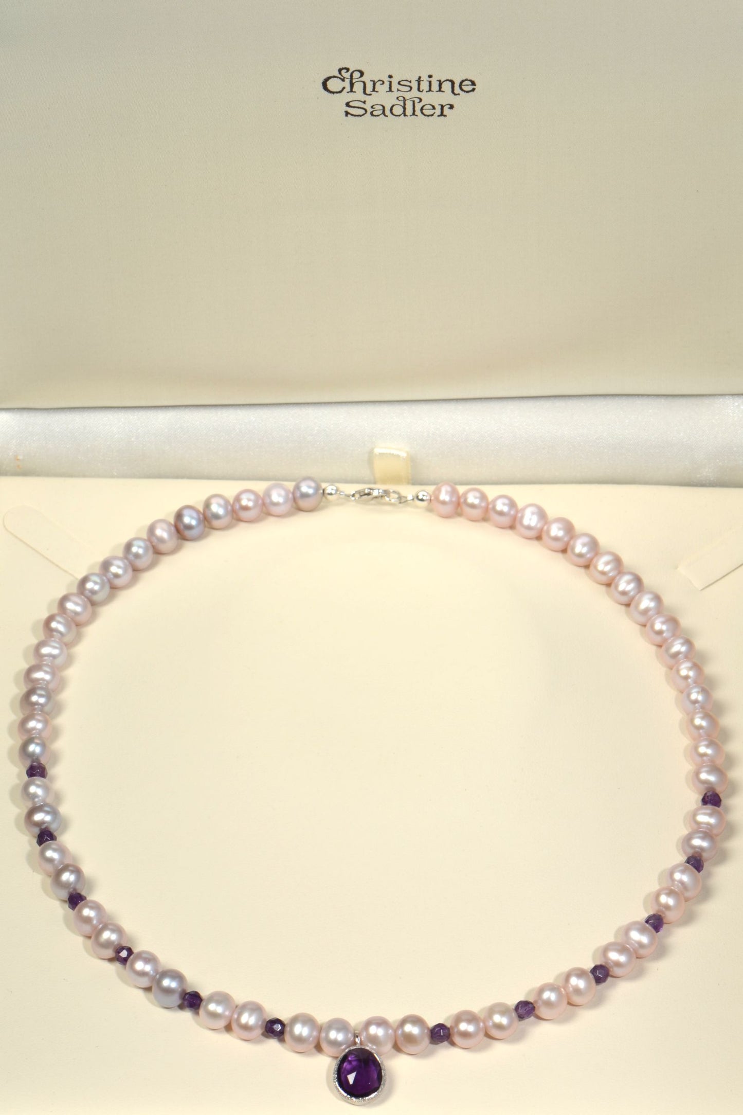 Pearl necklace with amethyst gemstone on a cream background, featuring the brand 'Christine Sadler'.