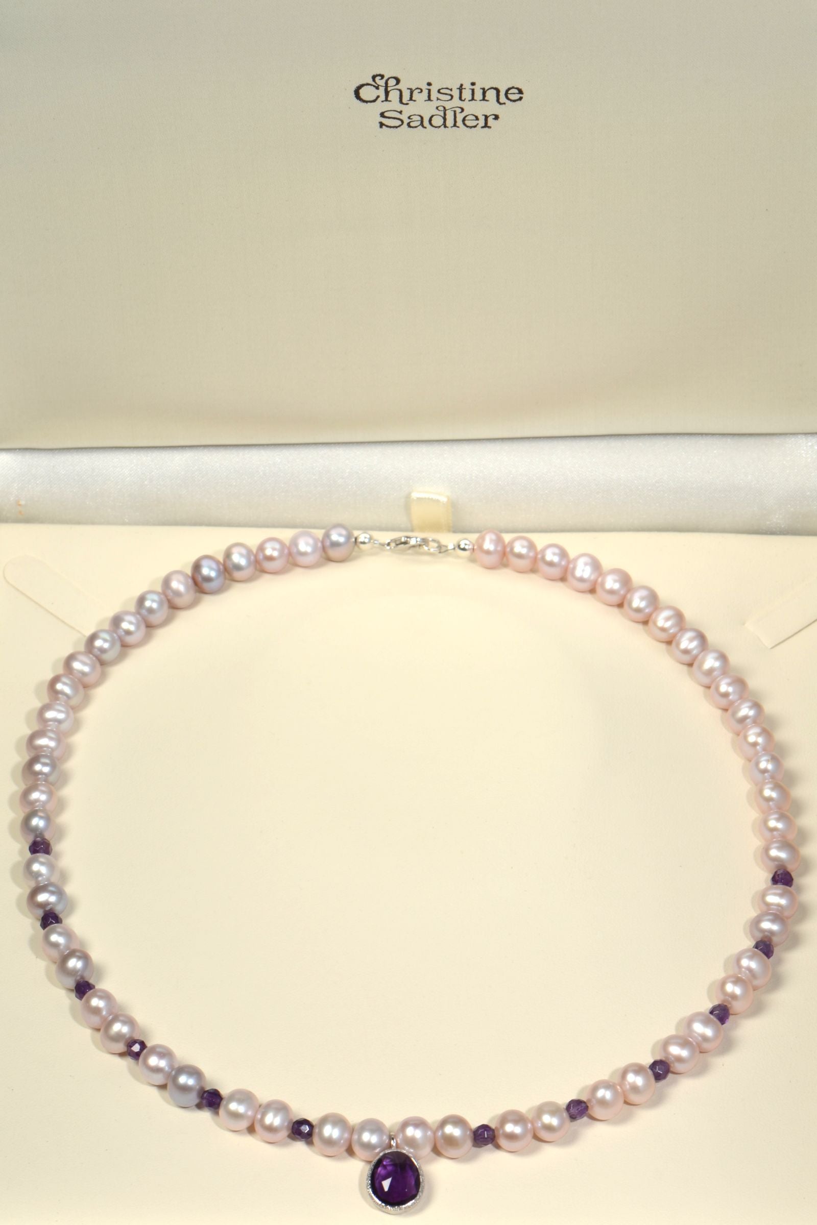 Pearl necklace with amethyst gemstone on a cream background, featuring the brand 'Christine Sadler'.