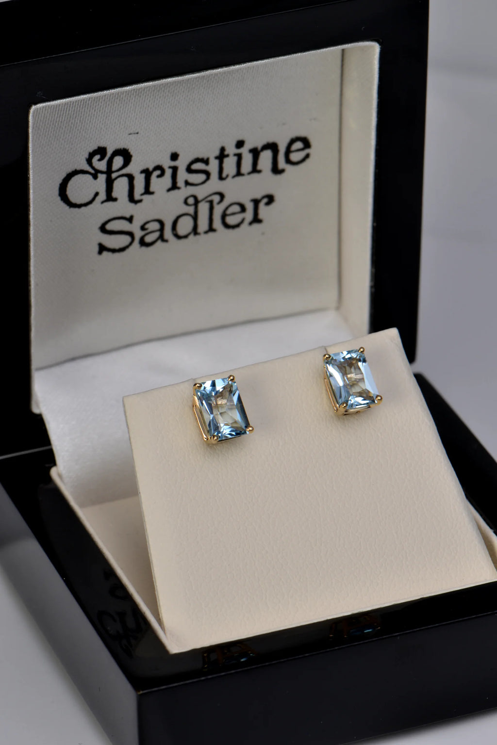 A pair of Christine Sadler stud earrings with rectangular aquamarine gemstones in gold settings, displayed on a cream jewellery stand inside an open black jewellery box.