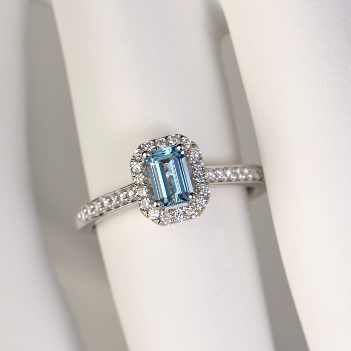 platinum halo ring set with an emerald cut aquamarine surrounded by small white round diamonds and diamonds in the band - engagement ring commission from Christine Sadler