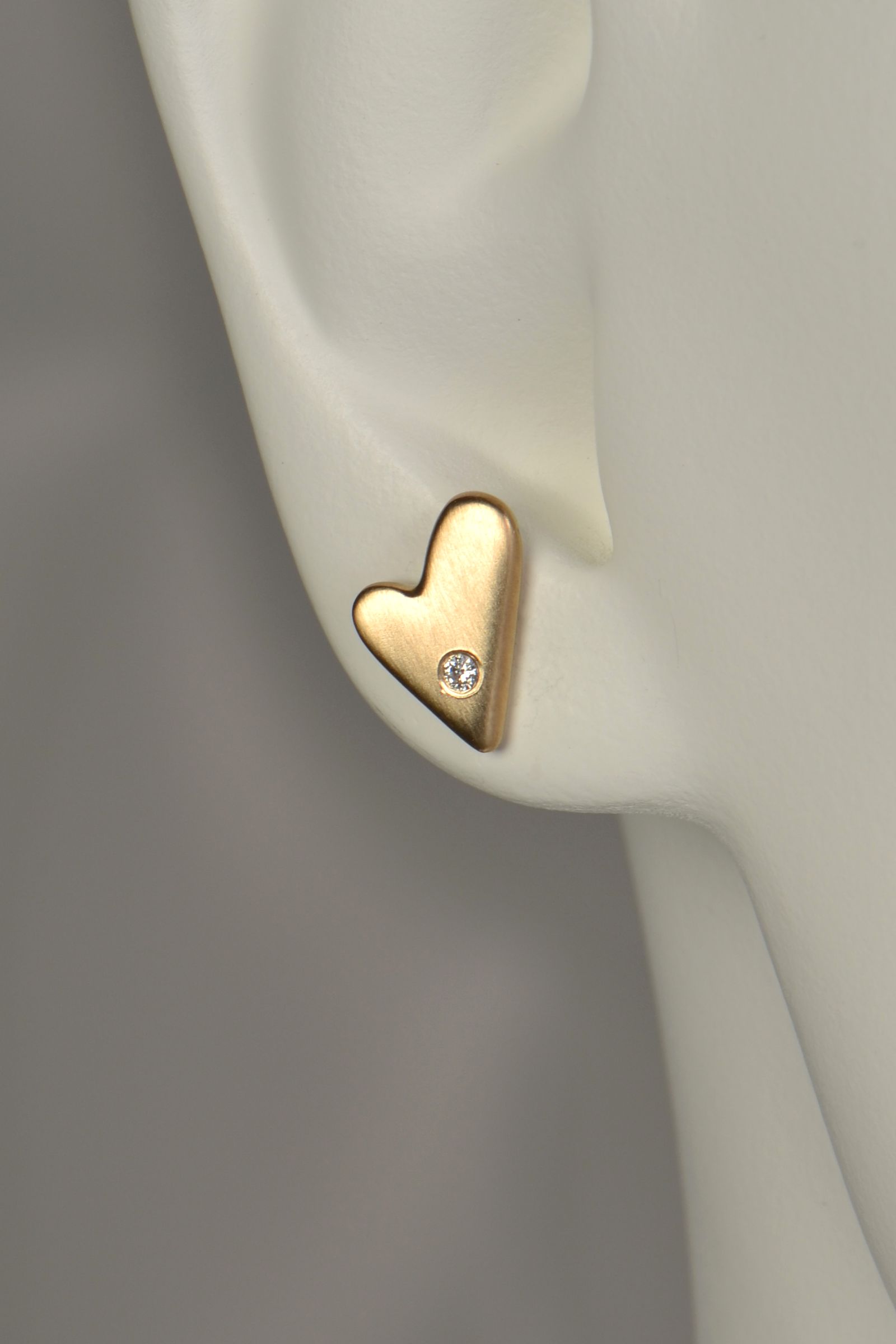 Gold heart-shaped earring on a mannequin ear against a gray background to show size and scale