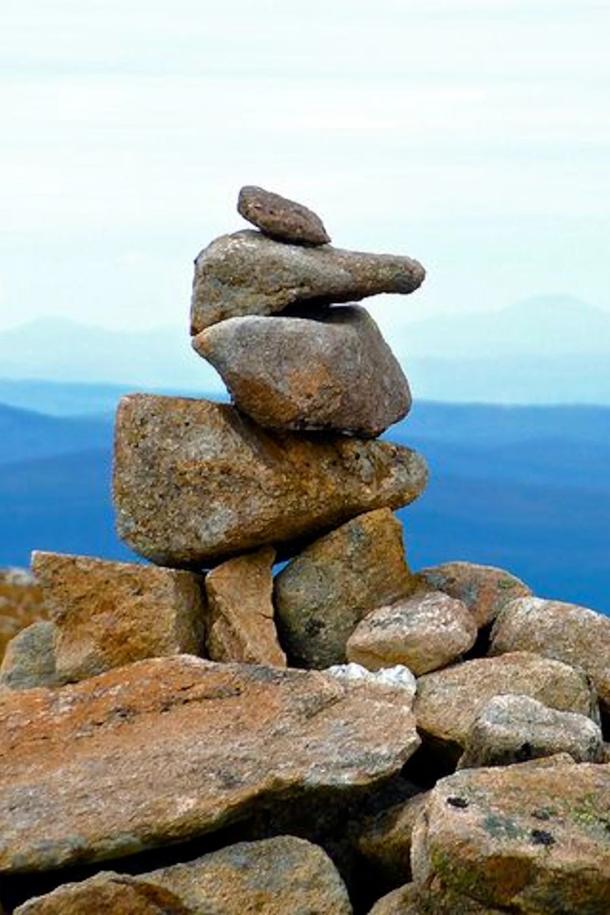 A balanced stack of stacked rocks (cairn) on a mountain peak with blue mountain ranges in the background under a pale sky showing where the inspiration for the curve design came from.