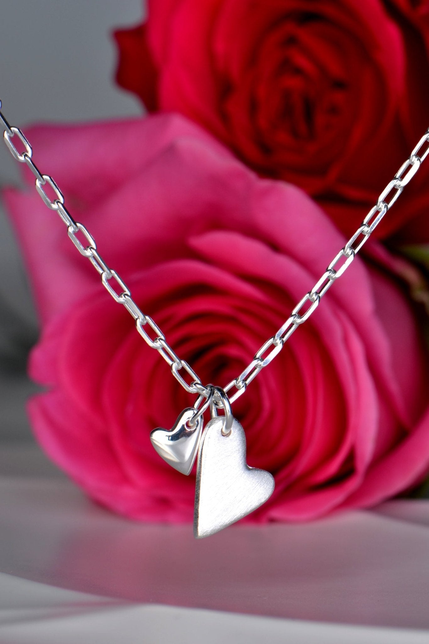 Sterling silver necklace with two heart pendants — one large matt heart and one small polished heart, From The Heart collection wiht red and pink roses in the background