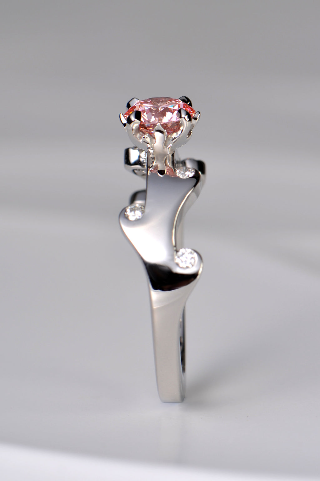 A stunning platinum engagement ring that is a unique design inspired by fire. The band waves in curving flames that hold small white lab diamonds and the main central round pink lab diamond is held by claws that are shaped into tiny flames.