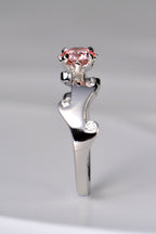 A stunning platinum engagement ring that is a unique design inspired by fire. The band waves in curving flames that hold small white lab diamonds and the main central round pink lab diamond is held by claws that are shaped into tiny flames.