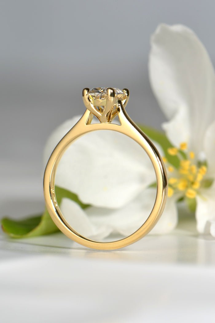 side view of an 18ct yellow gold engagement ring with an oval cut lab diamond and awhite flower that has yellow stamen and a green leaf in the background