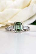 A 1.83ct trilogy ring crafted in platinum featuring a central 1.50ct emerald cut fancy vivid green lab-grown diamond held by four prongs and flanked by two tapered baguette side stones against a soft white rose background.