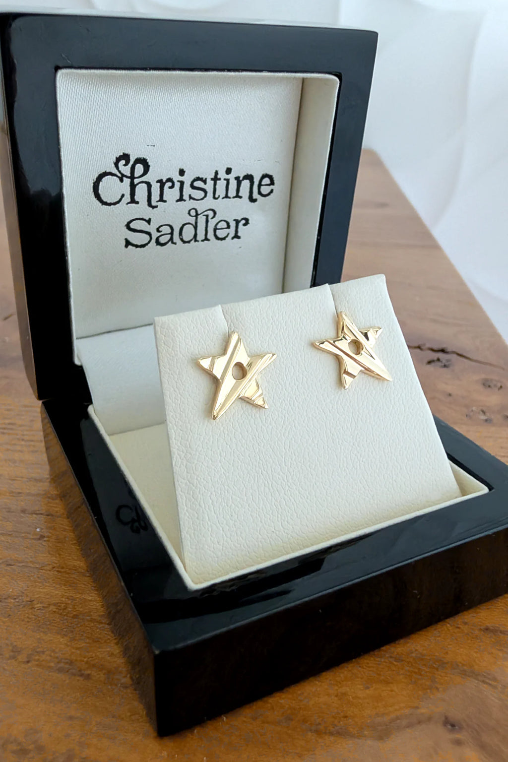 A pair of gold star-shaped stud earrings displayed on a cream jewelry stand inside an open black presentation box. The inside lid is printed with the brand name 'Christine Sadler'.