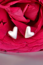 Close-up of a pair of handmade silver heart earrings – flat-edged, asymmetrical design with brushed finish. Pink rose in th background.