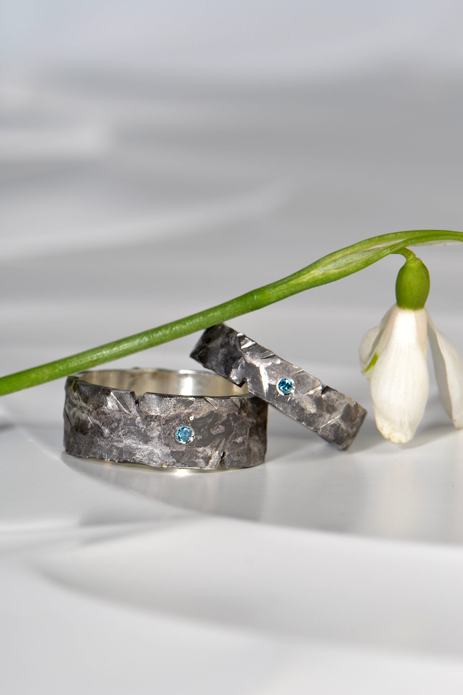 Two silver oxidised and textured rings with embedded blue diamonds on a white surface with a flower in the background from Christine Sadler's Hero collection where each piece is unique and one-of-a-kind