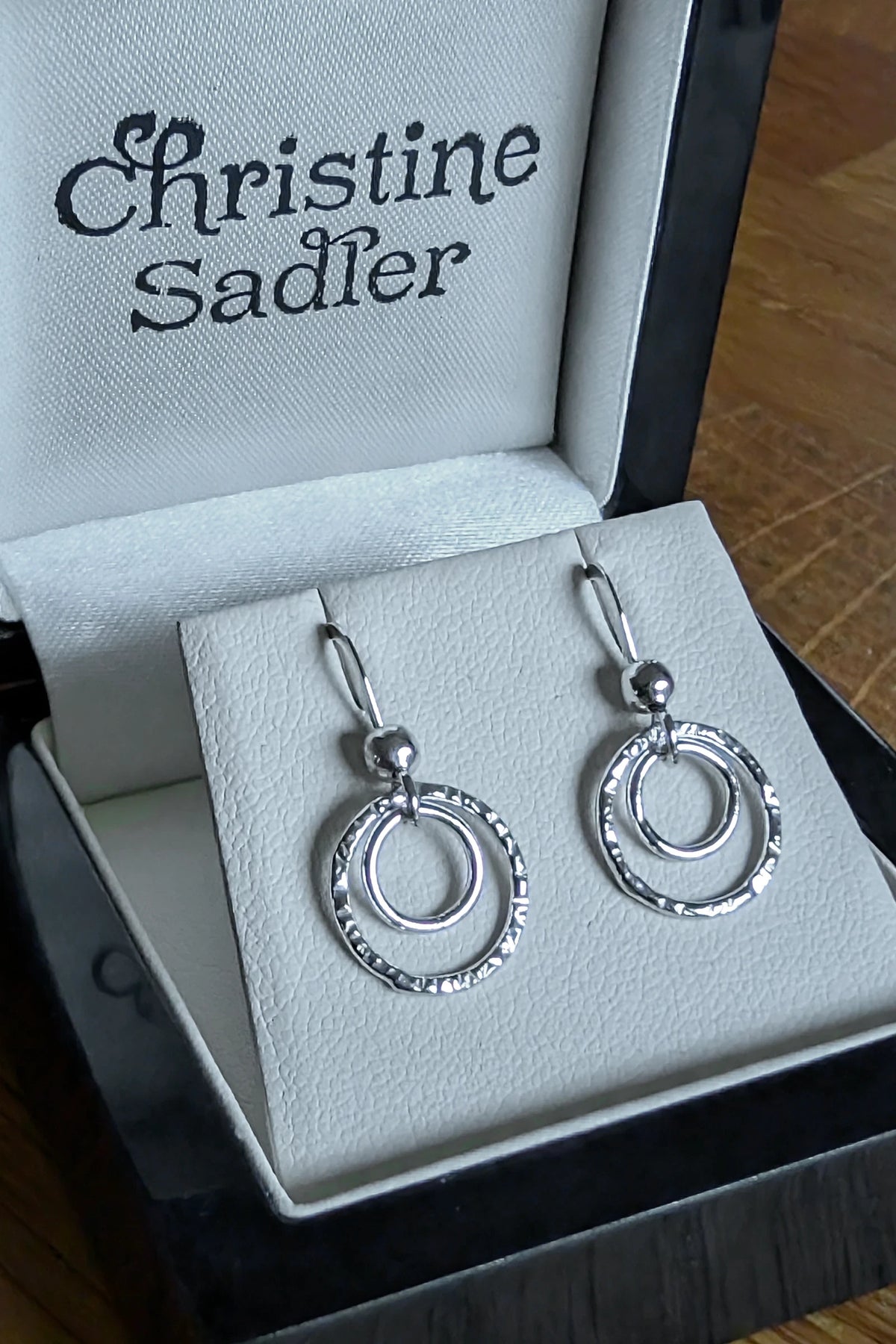 A pair of sterling silver double-ring dangle earrings with hook fittings inside an open black and cream Christine Sadler gift box, with the brand name printed on the inner lid.