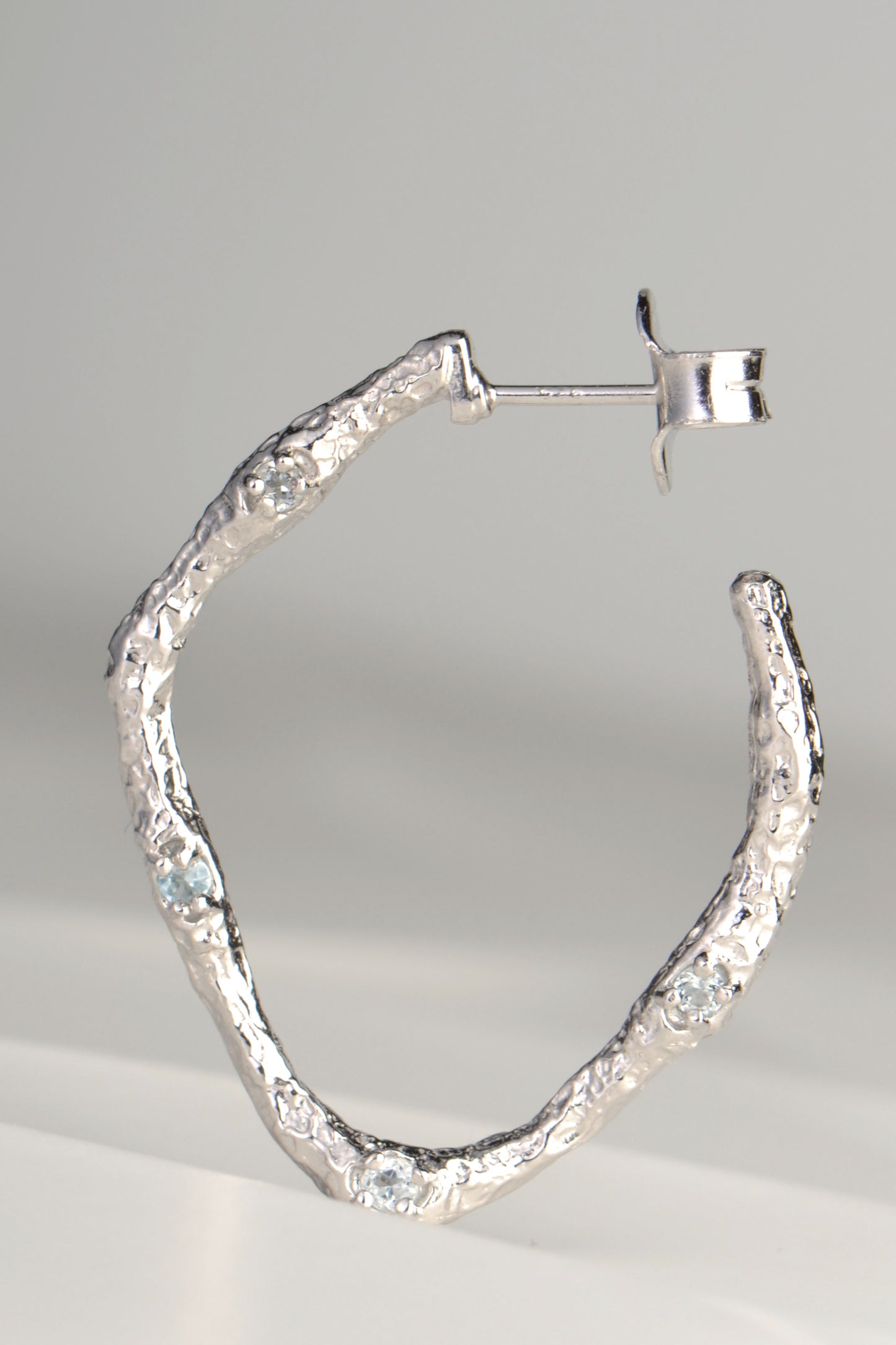 large silver hoop earrings with molton lava texture set with four very small pale blue topaz gemstones on one side by jewellery designer Jorge Revilla