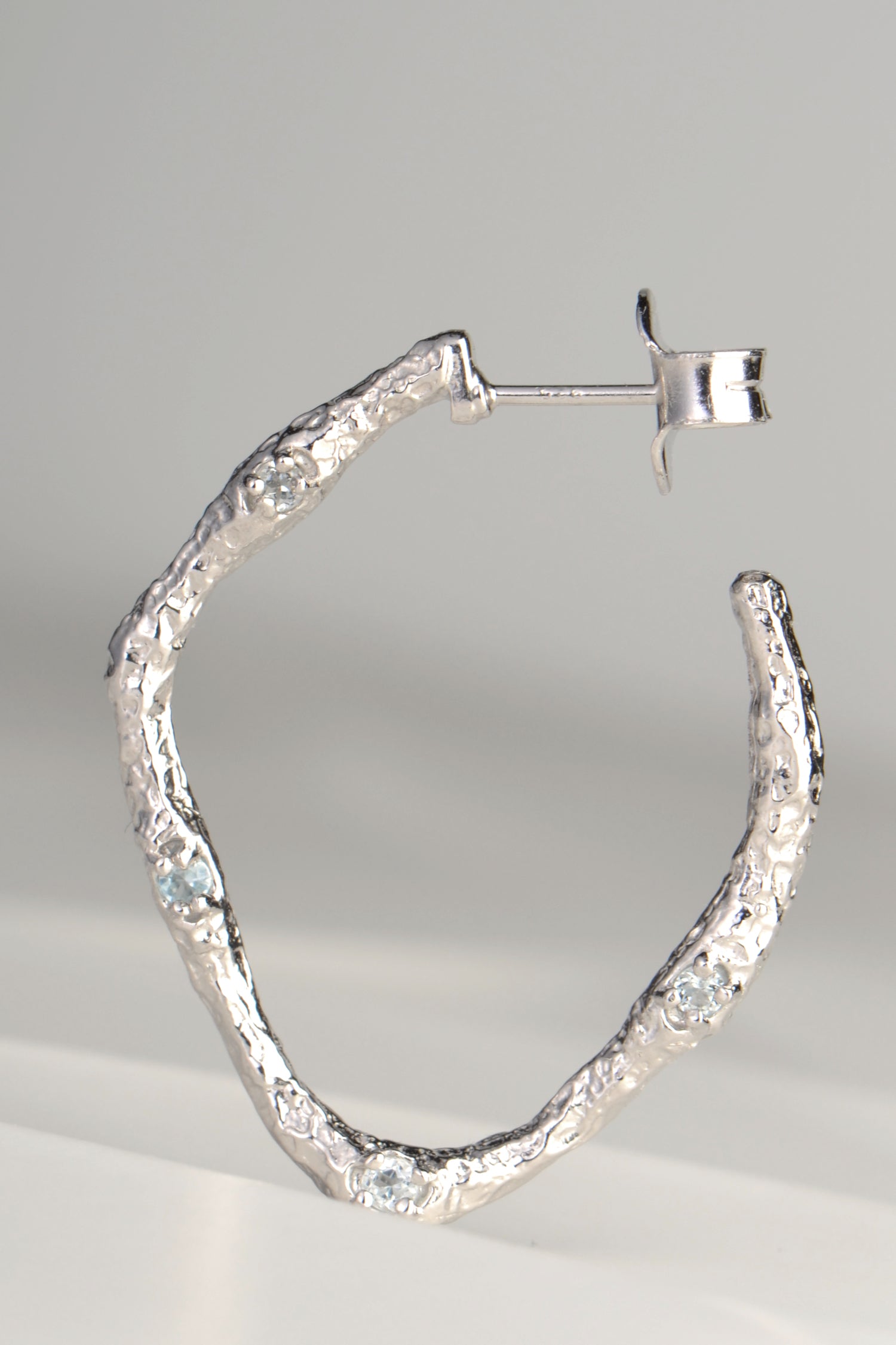 large silver hoop earrings with molton lava texture set with four very small pale blue topaz gemstones on one side by jewellery designer Jorge Revilla