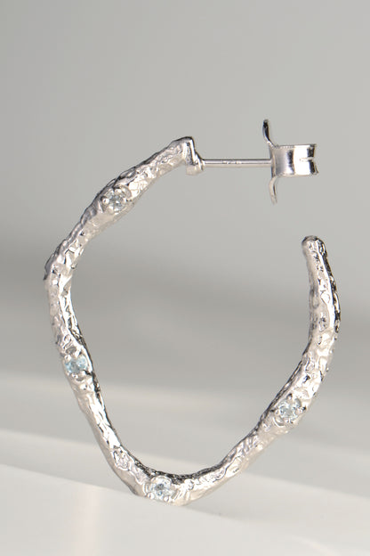 large silver hoop earrings with molton lava texture set with four very small pale blue topaz gemstones on one side by jewellery designer Jorge Revilla