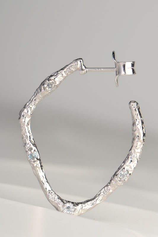 large silver hoop earrings with molton lava texture set with four very small pale blue topaz gemstones on one side by jewellery designer Jorge Revilla