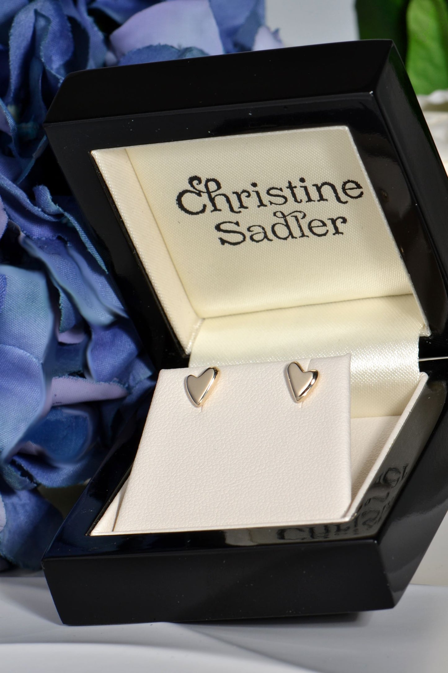 Heart-shaped gold earrings in a box with 'Christine Sadler' branding, surrounded by blue flowers.
