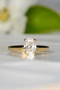 oval cut lab diamond engagement ring with a gold band. A white flower with a green leaf is blurred out in the background