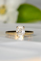 oval cut lab diamond engagement ring with a gold band. A white flower with a green leaf is blurred out in the background