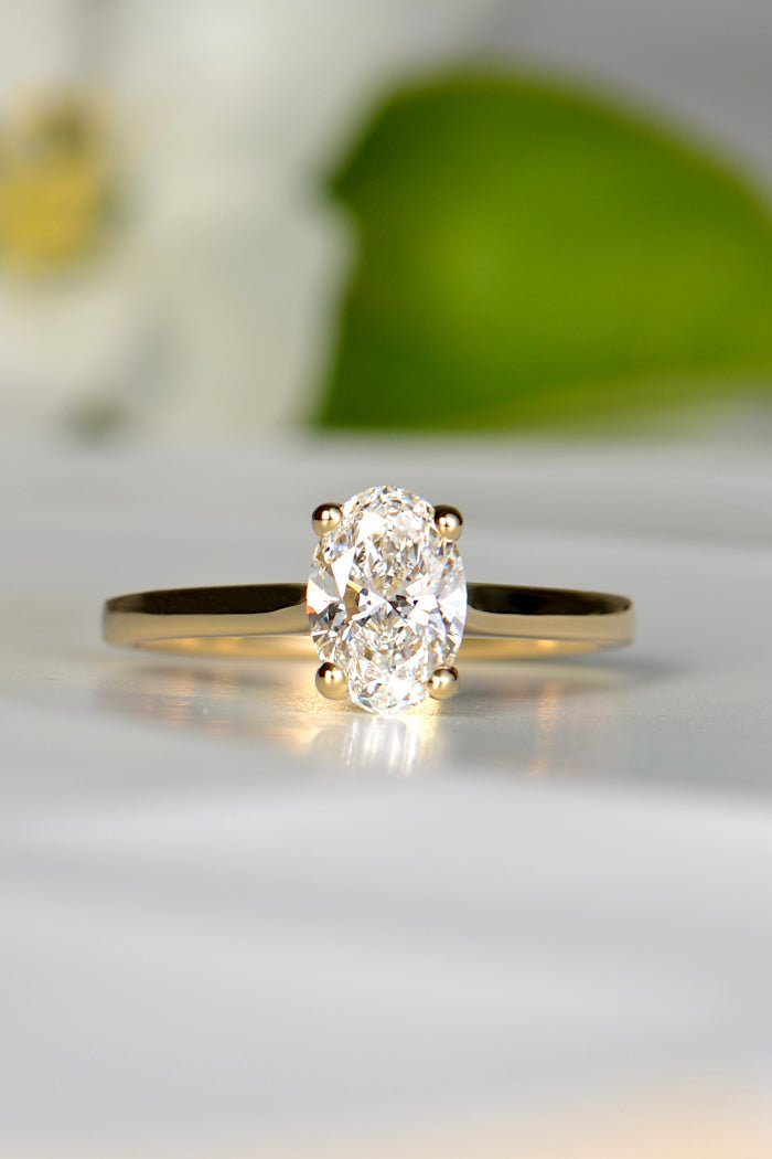 oval cut lab diamond engagement ring with a gold band. A white flower with a green leaf is blurred out in the background