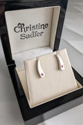 A pair of sterling silver curved stud earrings with small ruby gemstones, displayed on a cream jewellery stand inside an open black wooden Christine Sadler gift box.