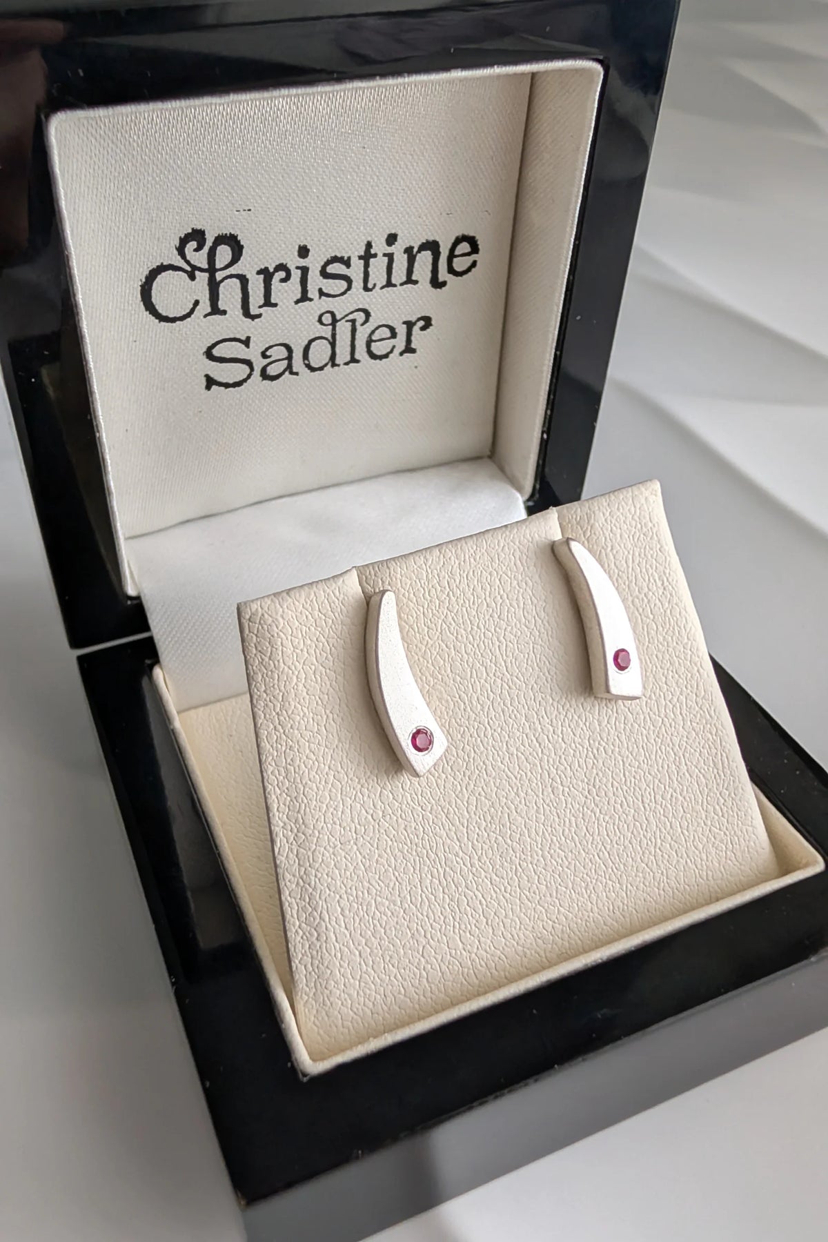 A pair of sterling silver curved stud earrings with small ruby gemstones, displayed on a cream jewellery stand inside an open black wooden Christine Sadler gift box.