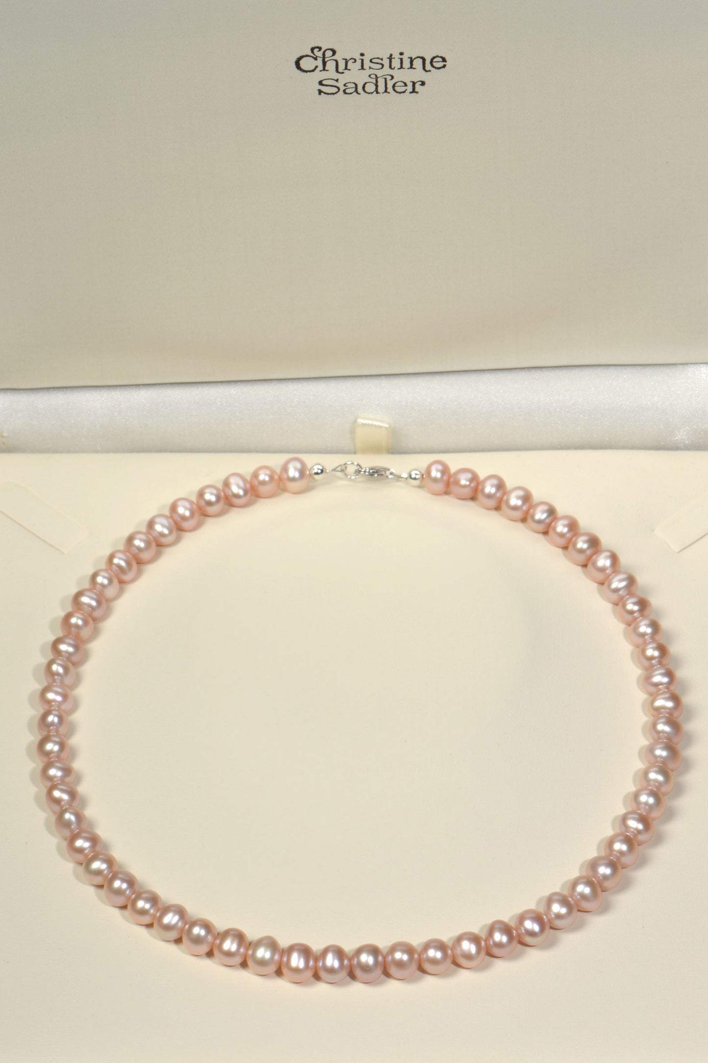 Pink cultured pearl necklace on a cream presentation box with 'Christine Sadler' branding.