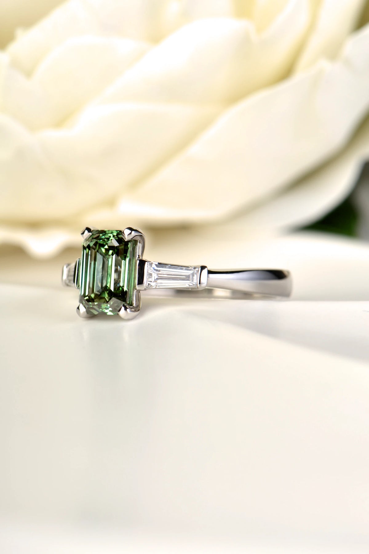 An angled close-up view of the 1.83ct platinum trilogy ring on a white rose petal, highlighting the structural durability, secure prong placement, and vivid color of the central green lab-grown diamond.