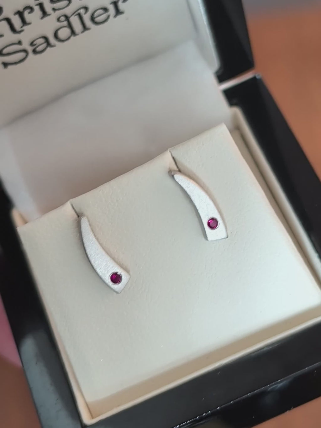 Tapered curve brushed sterling silver handmade earrings set with a real ruby at the bottom of each shown in the box in natural daylight and rotated to show the high quality post and butterfly fittings.