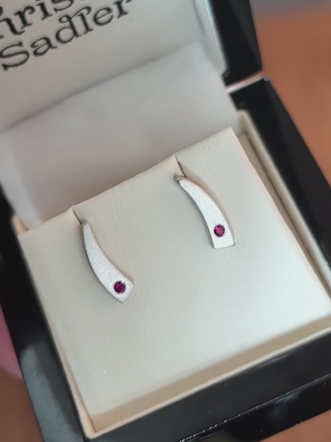 Tapered curve brushed sterling silver handmade earrings set with a real ruby at the bottom of each shown in the box in natural daylight and rotated to show the high quality post and butterfly fittings.