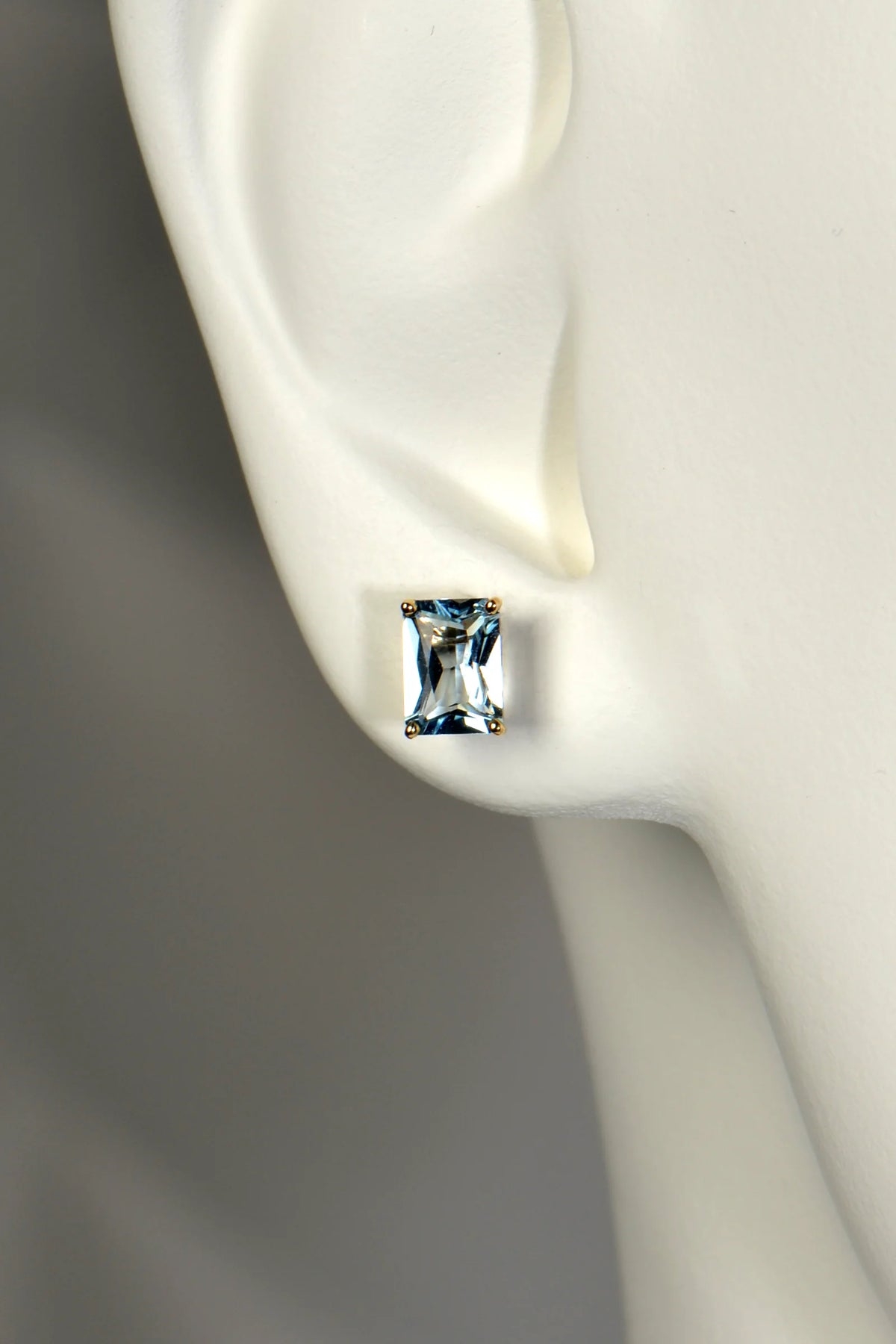 A rectangular aquamarine gemstone stud earring with 9ct yellow gold prongs, shown on a white mannequin ear against a soft gray background.