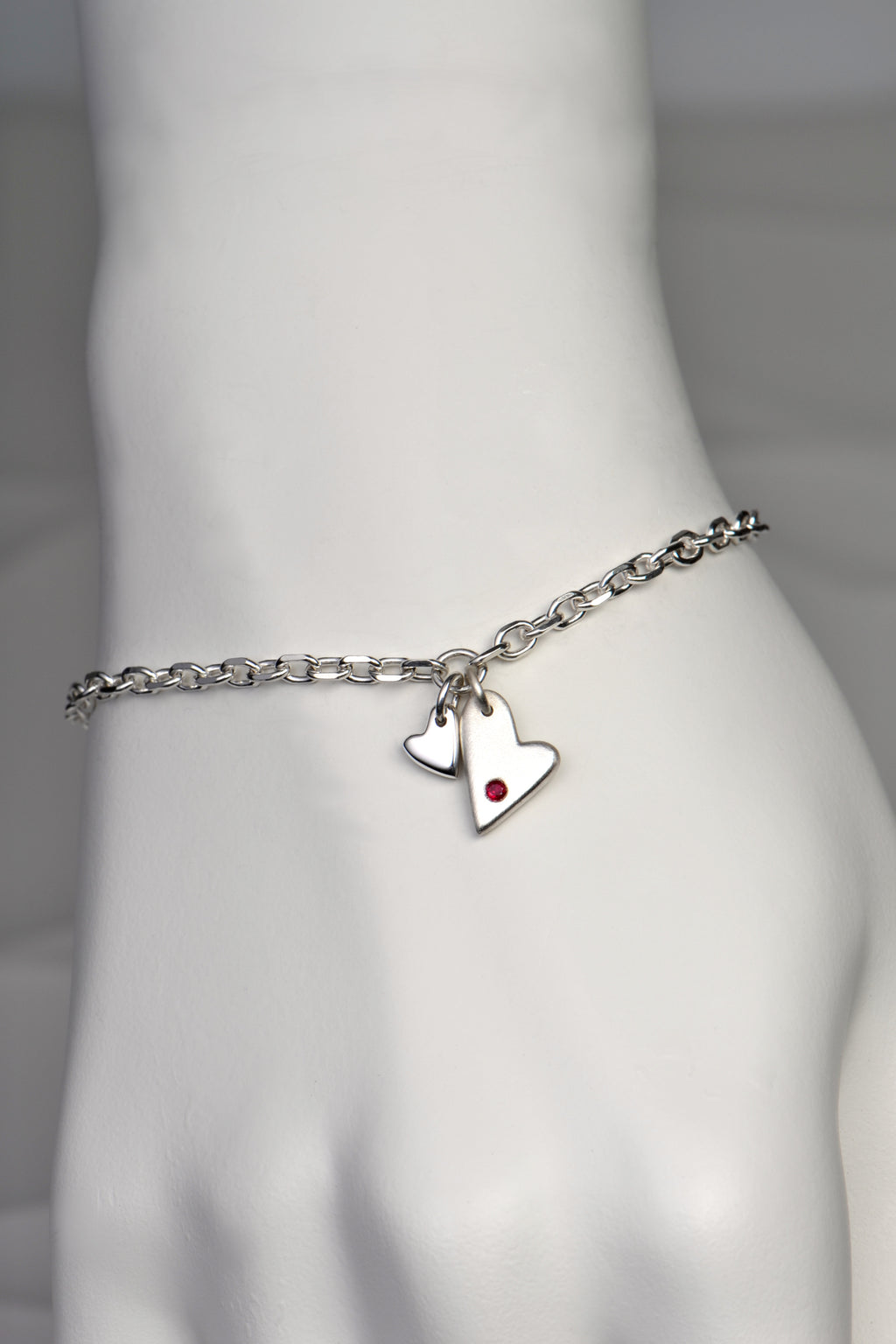 handmade silver heart bracelet by Christine Sadler. Two hearts hang at the front, one larger than the other. The larger heart is matt finished and set with a ruby gemstone