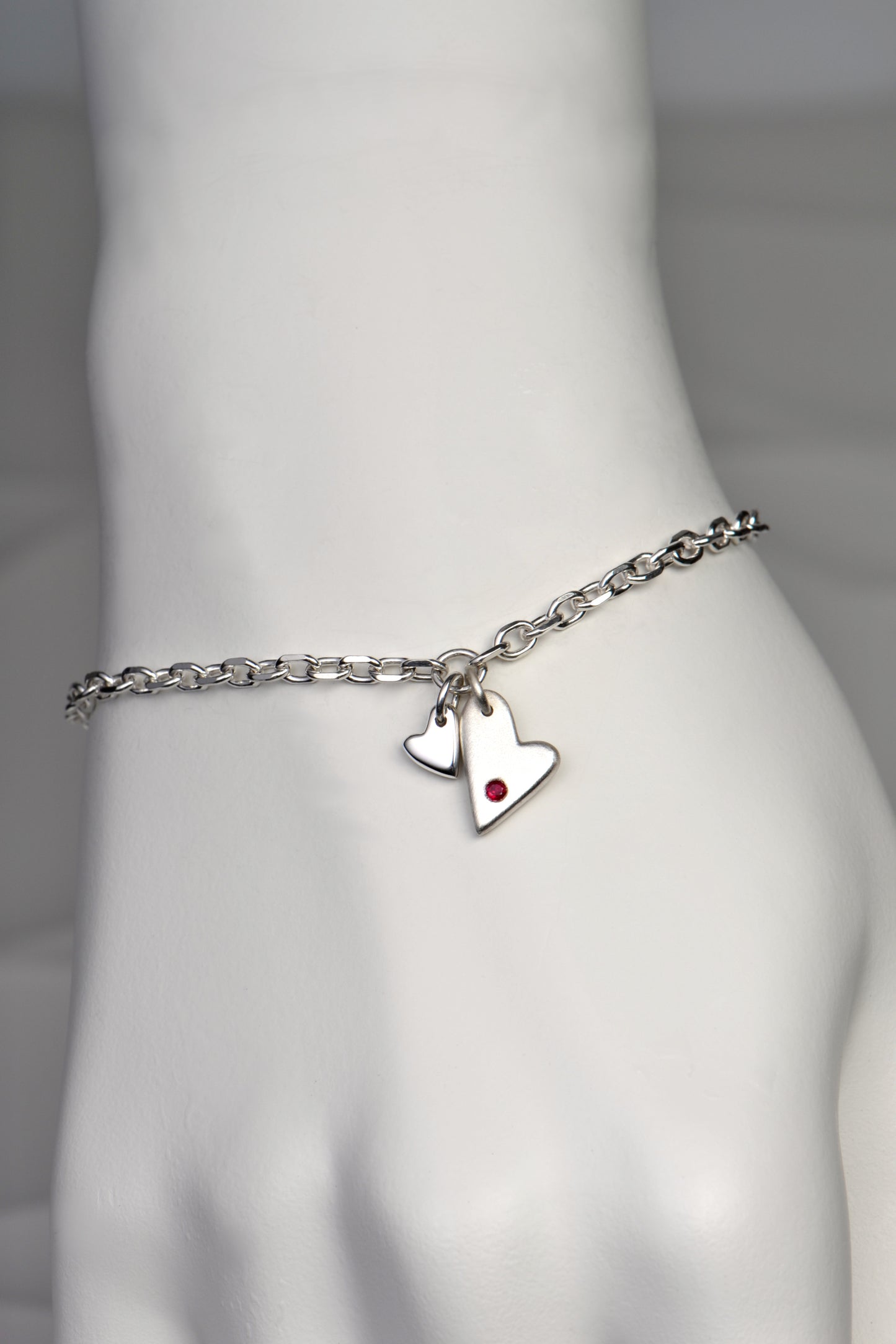 handmade silver heart bracelet by Christine Sadler. Two hearts hang at the front, one larger than the other. The larger heart is matt finished and set with a ruby gemstone