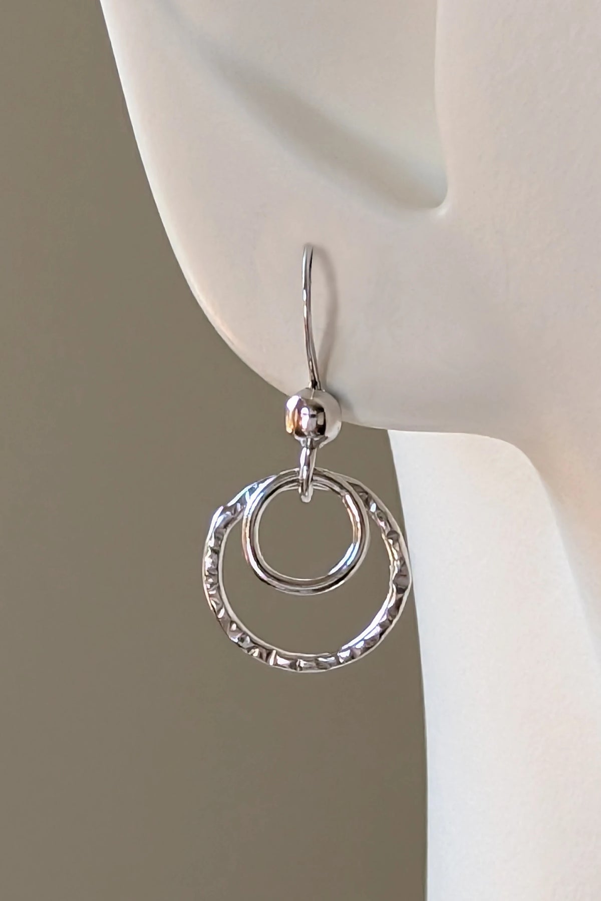 A sterling silver double-ring dangle earrings displayed on a white mannequin ear against a lichen green background.