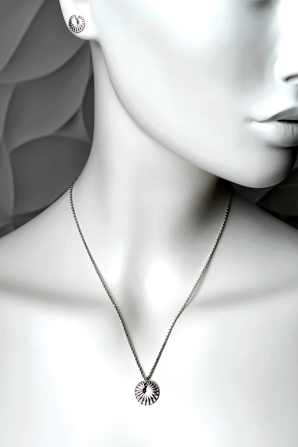 A sterling silver circular pendant necklace with a spoked wheel design, engraved "enough" displayed on a white mannequin bust. The mannequin also wears a matching round stud earring.