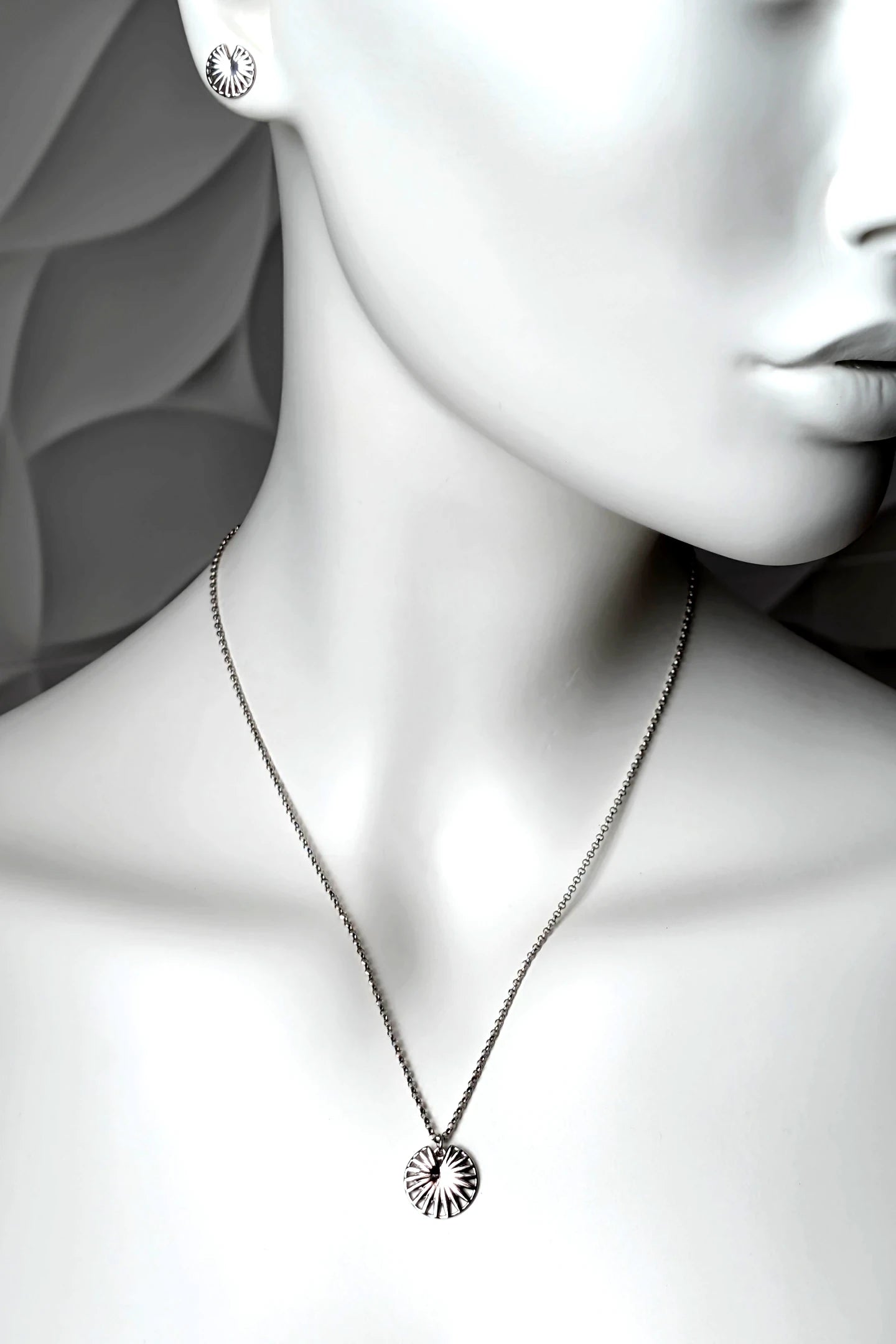 A sterling silver circular pendant necklace with a spoked wheel design, engraved "enough" displayed on a white mannequin bust. The mannequin also wears a matching round stud earring.