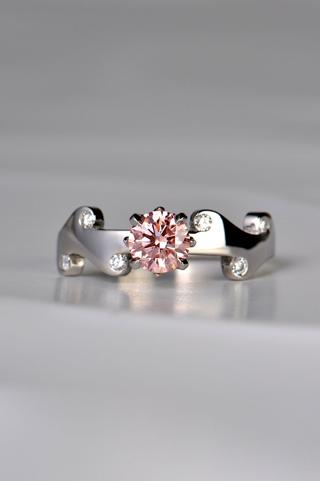 Large round pink lab diamond set in the middle of a platinum wavy ring with 6 small round white diamonds scattered across the front half of the band. This unique design by Christine Sadler is inspired by fire and the pink lab diamond is held by claws that are shaped into tiny flames. 