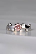 Large round pink lab diamond set in the middle of a platinum wavy ring with 6 small round white diamonds scattered across the front half of the band. This unique design by Christine Sadler is inspired by fire and the pink lab diamond is held by claws that are shaped into tiny flames. 