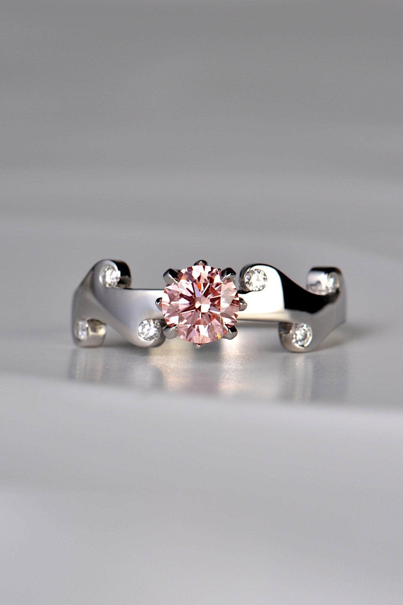 Large round pink lab diamond set in the middle of a platinum wavy ring with 6 small round white diamonds scattered across the front half of the band. This unique design by Christine Sadler is inspired by fire and the pink lab diamond is held by claws that are shaped into tiny flames. 