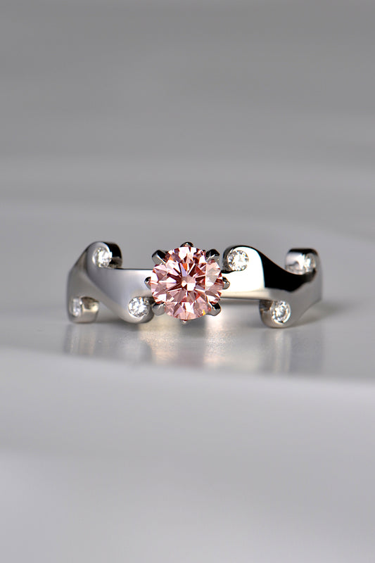 Large round pink lab diamond set in the middle of a platinum wavy ring with 6 small round white diamonds scattered across the front half of the band. This unique design by Christine Sadler is inspired by fire and the pink lab diamond is held by claws that are shaped into tiny flames. 