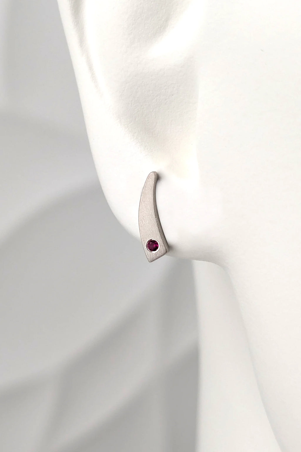 A handmade matt finished small silver crescent-shaped stud earring with a round ruby gemstone, displayed on a white mannequin ear for size and scale. 